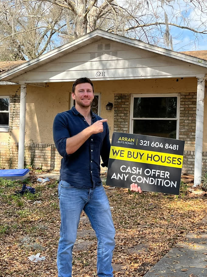 Arran holding an Arran Buys Homes sign at a house needing repair