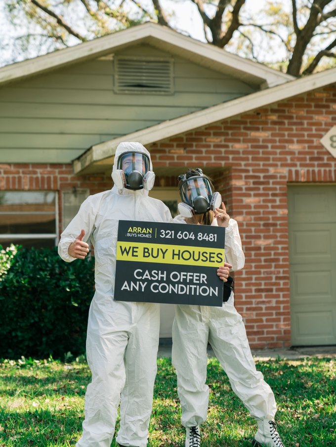 Arran and a crew member in protective suits and respirators holding a We Buy Houses sign in front of a home being cleaned out